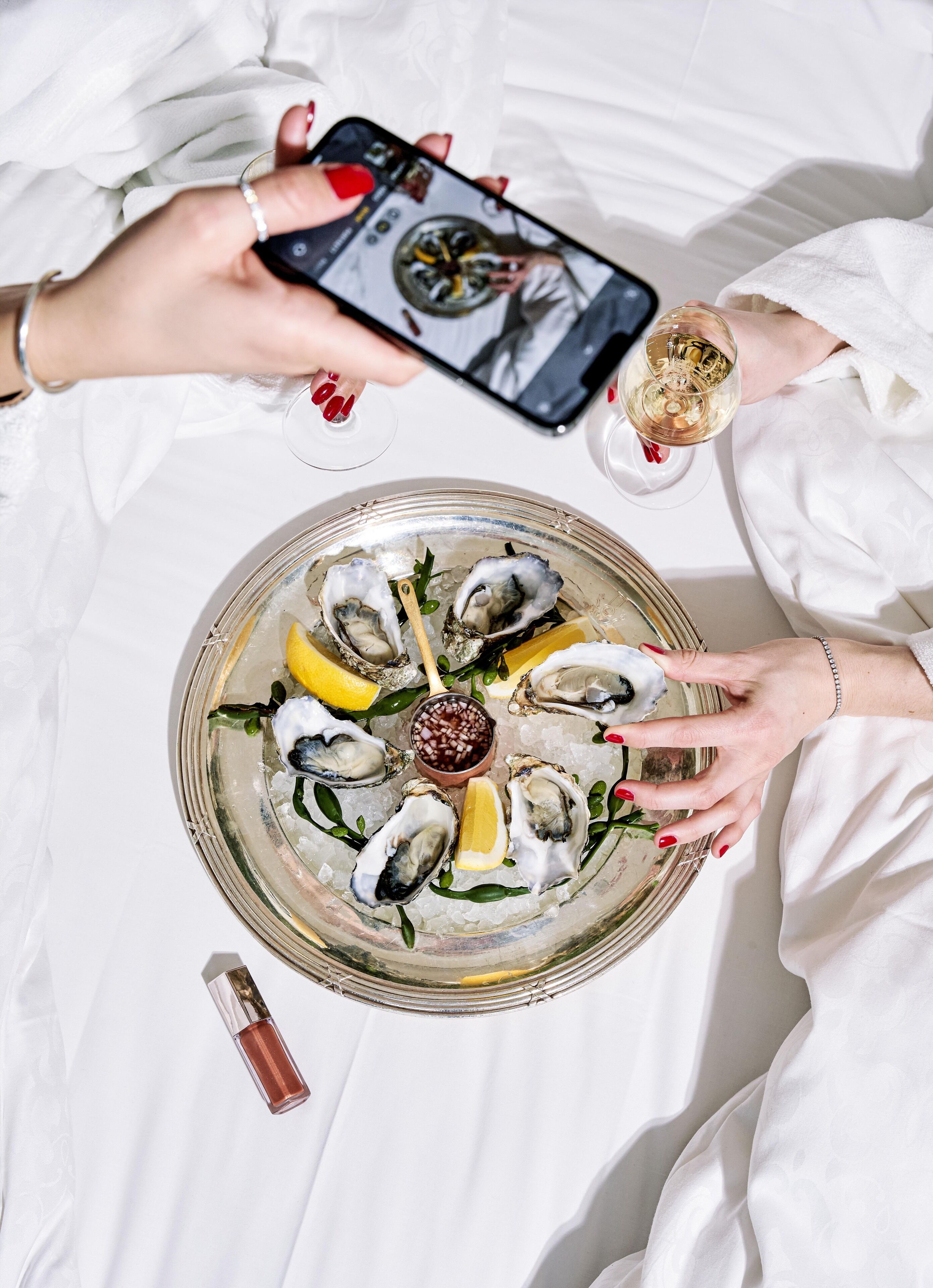 A tray of oysters with lemon wedges and a small bowl of sauce, a glass of white wine, and a smartphone being used to take a photo.