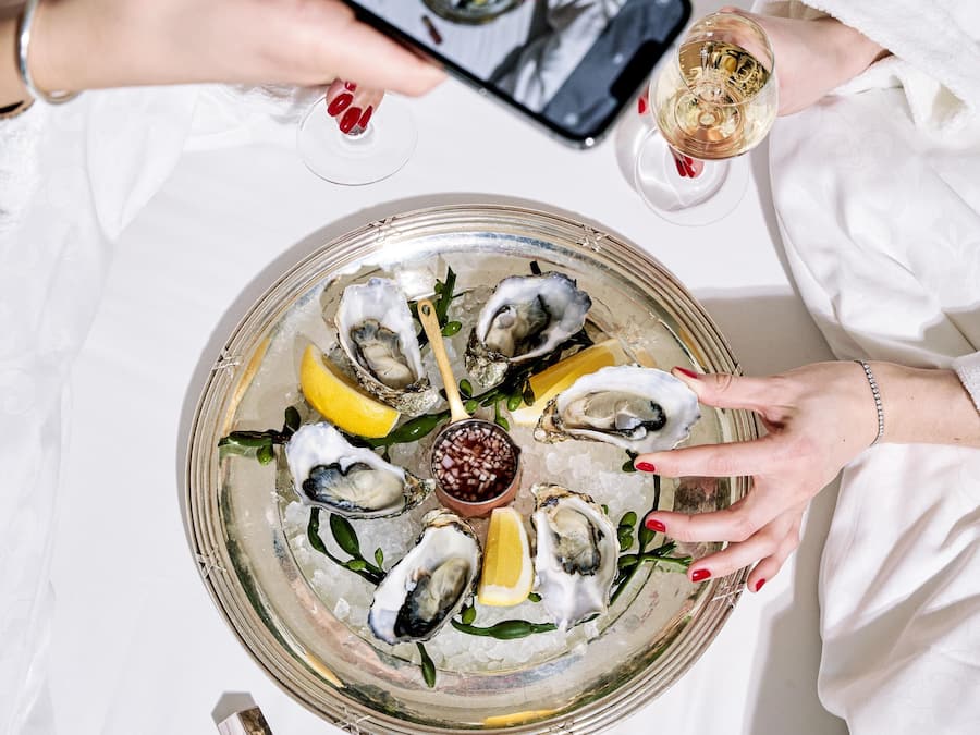 A tray of oysters with lemon wedges and a small bowl of sauce, a glass of white wine, and a smartphone being used to take a photo.