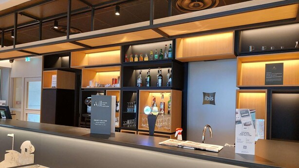 A hotel reception area with a counter, shelves with bottles, and a sign for 'Priority Welcome.' - ibis Besancon Centre Ville (Besancon)