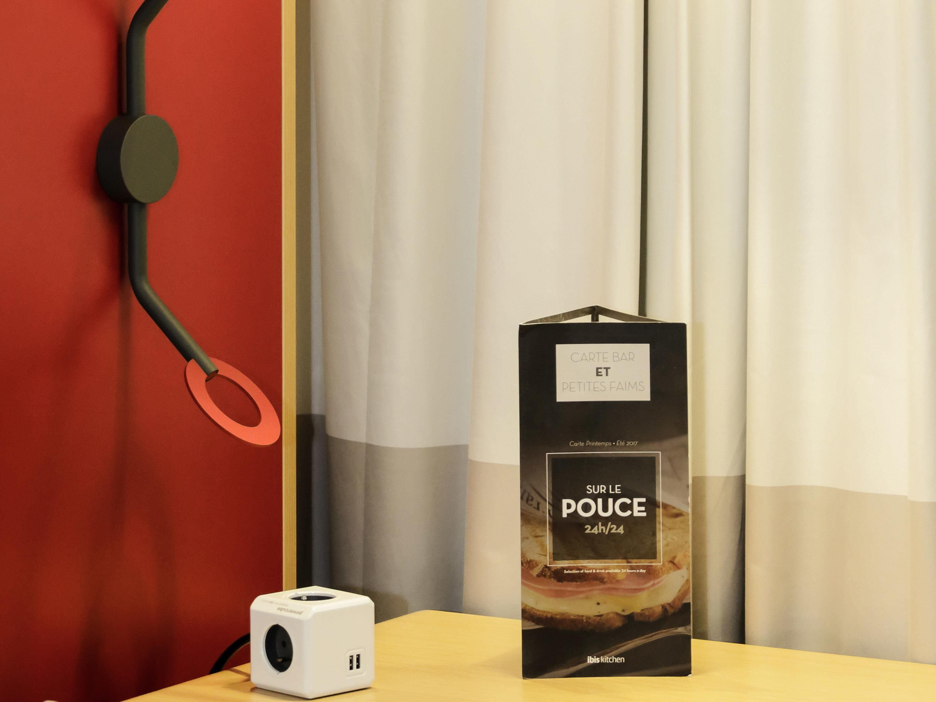 A white device on a wooden surface, a black box with text, and a red wall with a black object.