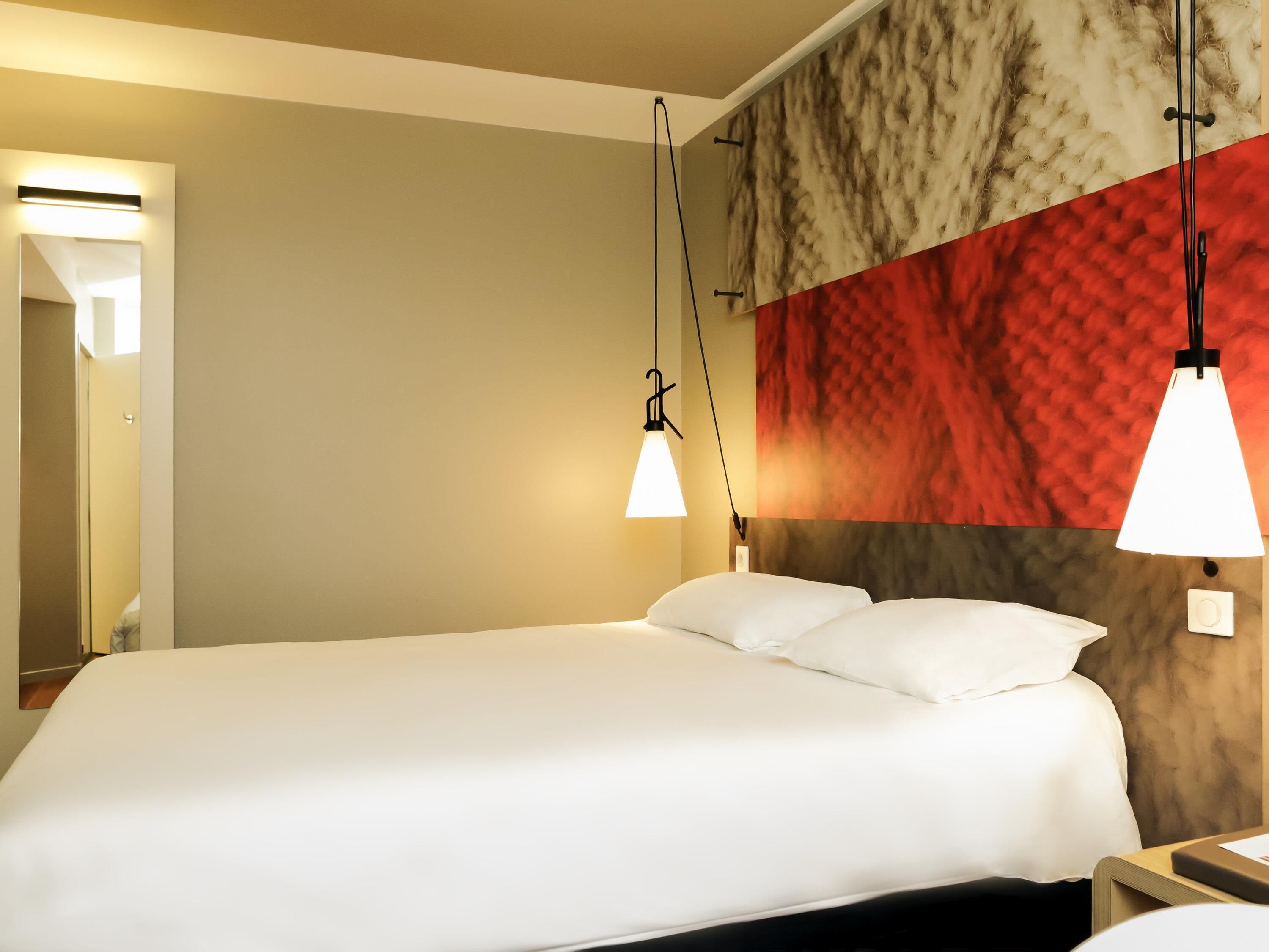A hotel room with a large bed, two bedside lamps, a wall-mounted mirror, and a red textured wall.
