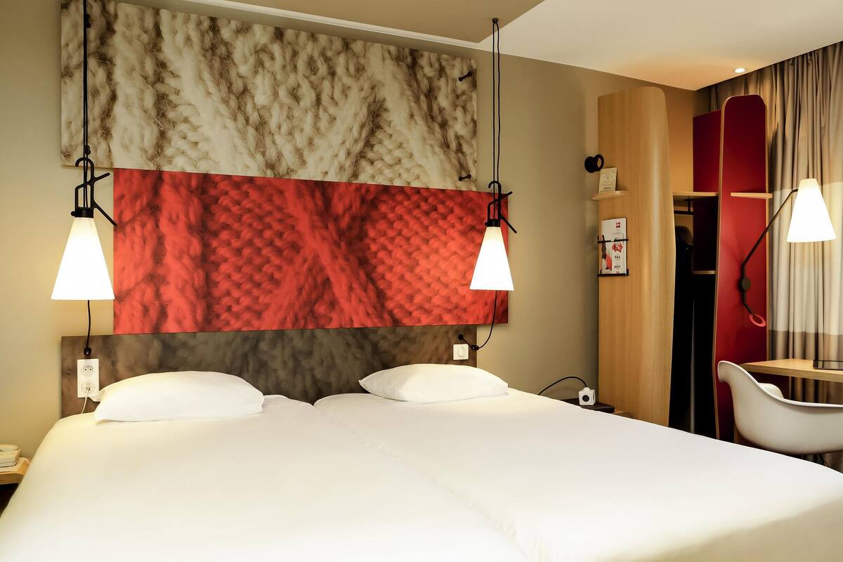 A hotel room with a bed, two bedside lamps, a red and white patterned headboard, a wooden wardrobe, a mirror, and a chair.