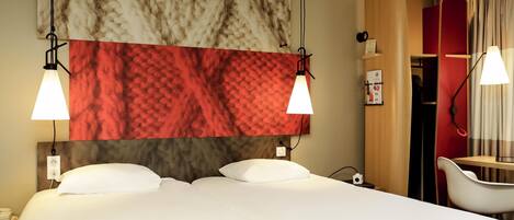 A hotel room with a bed, two bedside lamps, a red and white patterned headboard, a wooden wardrobe, a mirror, and a chair.