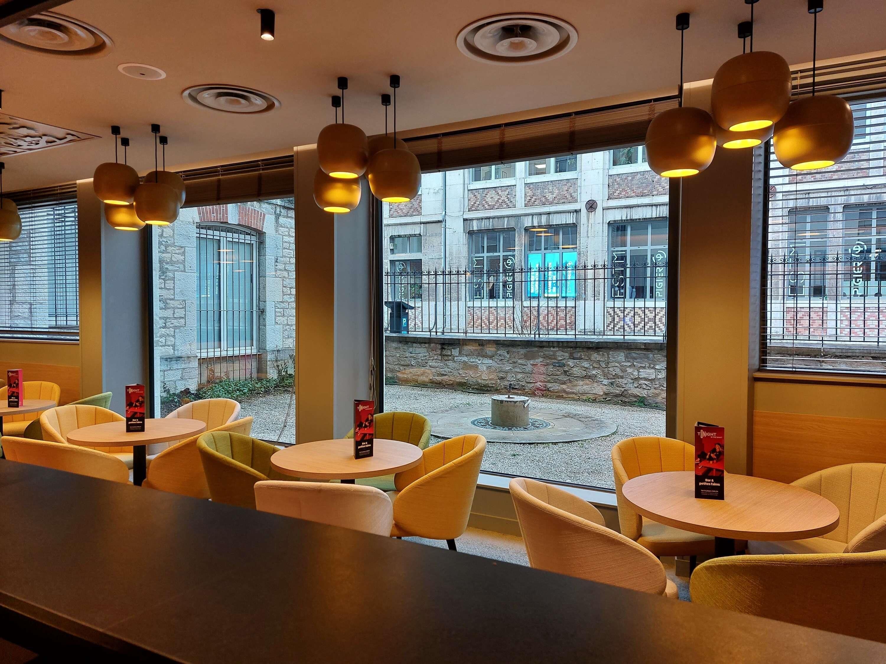 Een restaurantinterieur met ronde tafels en gele stoelen, grote ramen met uitzicht op de straat en hanglampen.