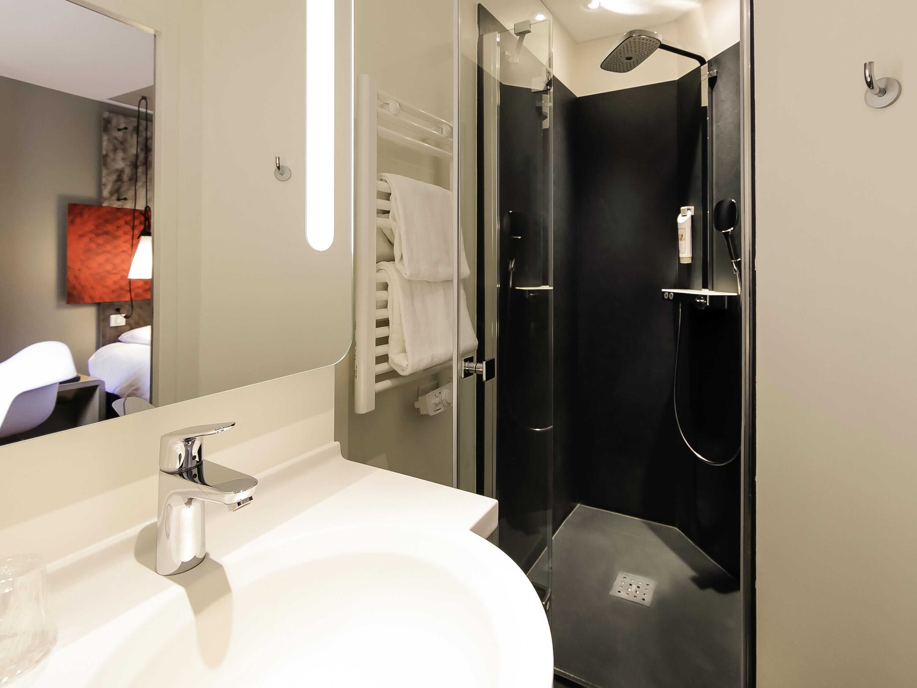 Casa de banho moderna com lavatório branco, cabine de duche em vidro e espelho a refletir uma cama e uma lâmpada fixada na parede.