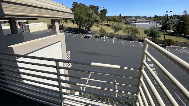 A parking lot with a few cars, a building with a beige roof, and trees in the background. - Budget Inn of Hayward (Hayward)