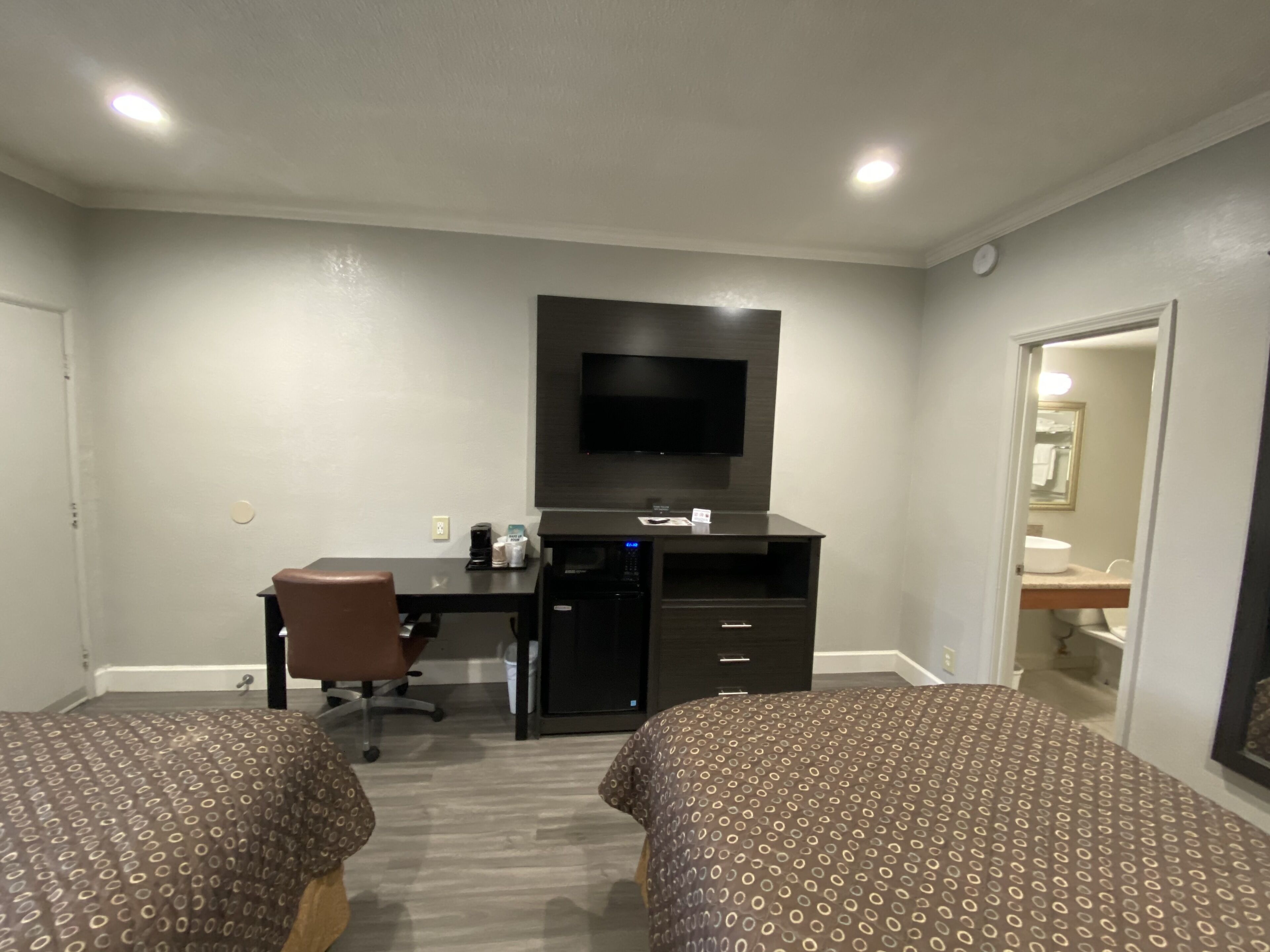 Ein Hotelzimmer mit zwei Betten, einem Schreibtisch mit Computer, einem an der Wand befestigten Fernseher und einem durchgehenden Tür zu einem Badezimmer.