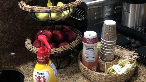 A kitchen counter with a basket of apples, a bottle of coffee-mate, a bottle of mustard, a coffee grinder, and a kettle. - Budget Inn of Hayward (Hayward)