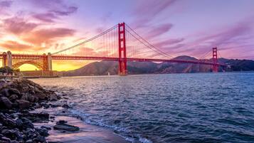 A suspension bridge with a prominent red structure, a rocky shoreline, and a sunset in the background.