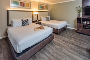 A hotel room with two beds, a TV, a bedside table with a lamp, and a small plant.
