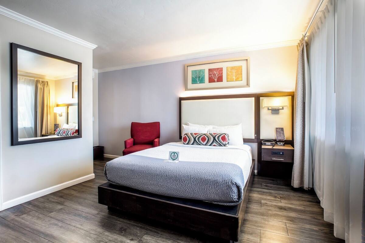 A hotel room with a large bed, a red chair, a nightstand, and a wall-mounted mirror.