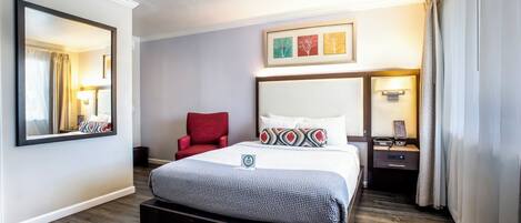 A hotel room with a large bed, a red chair, a nightstand, and a wall-mounted mirror.