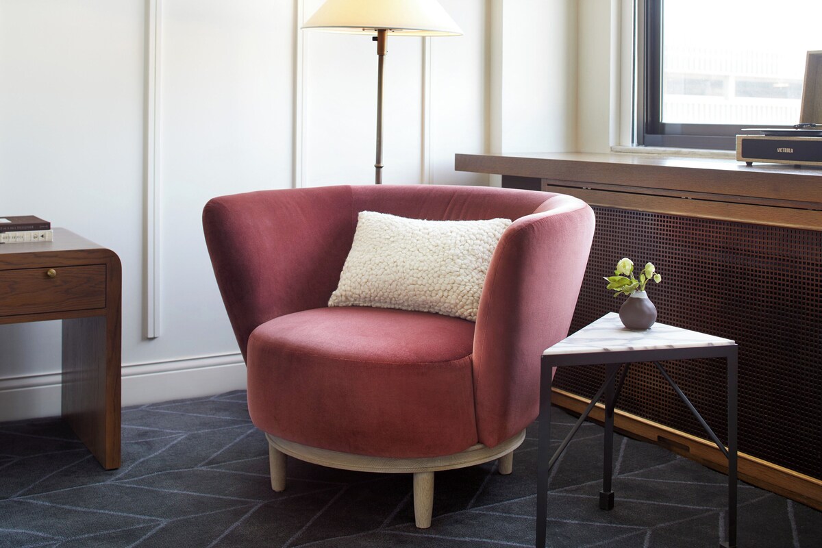 A red armchair with a white cushion, a small table with a vase, and a floor lamp in a room with a radiator and a window.