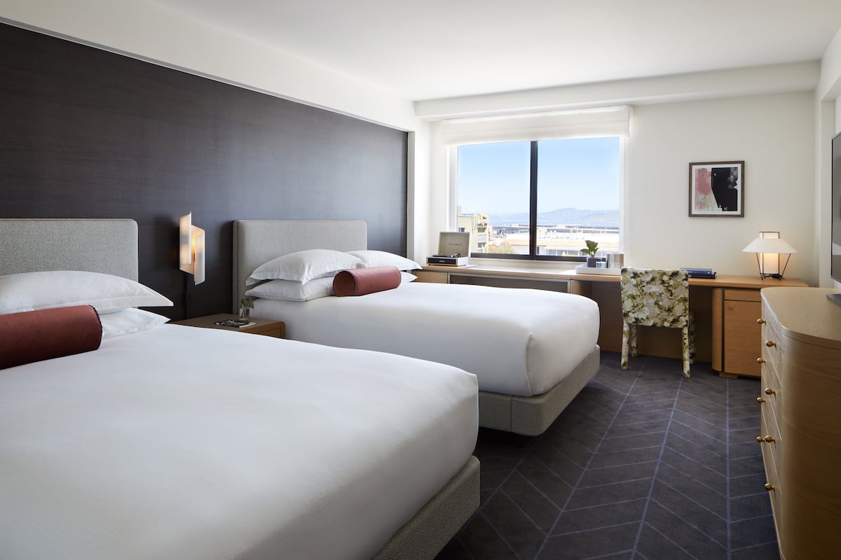 A hotel room with two beds, a desk, a chair, a dresser, a window with a view, and a wall-mounted lamp.