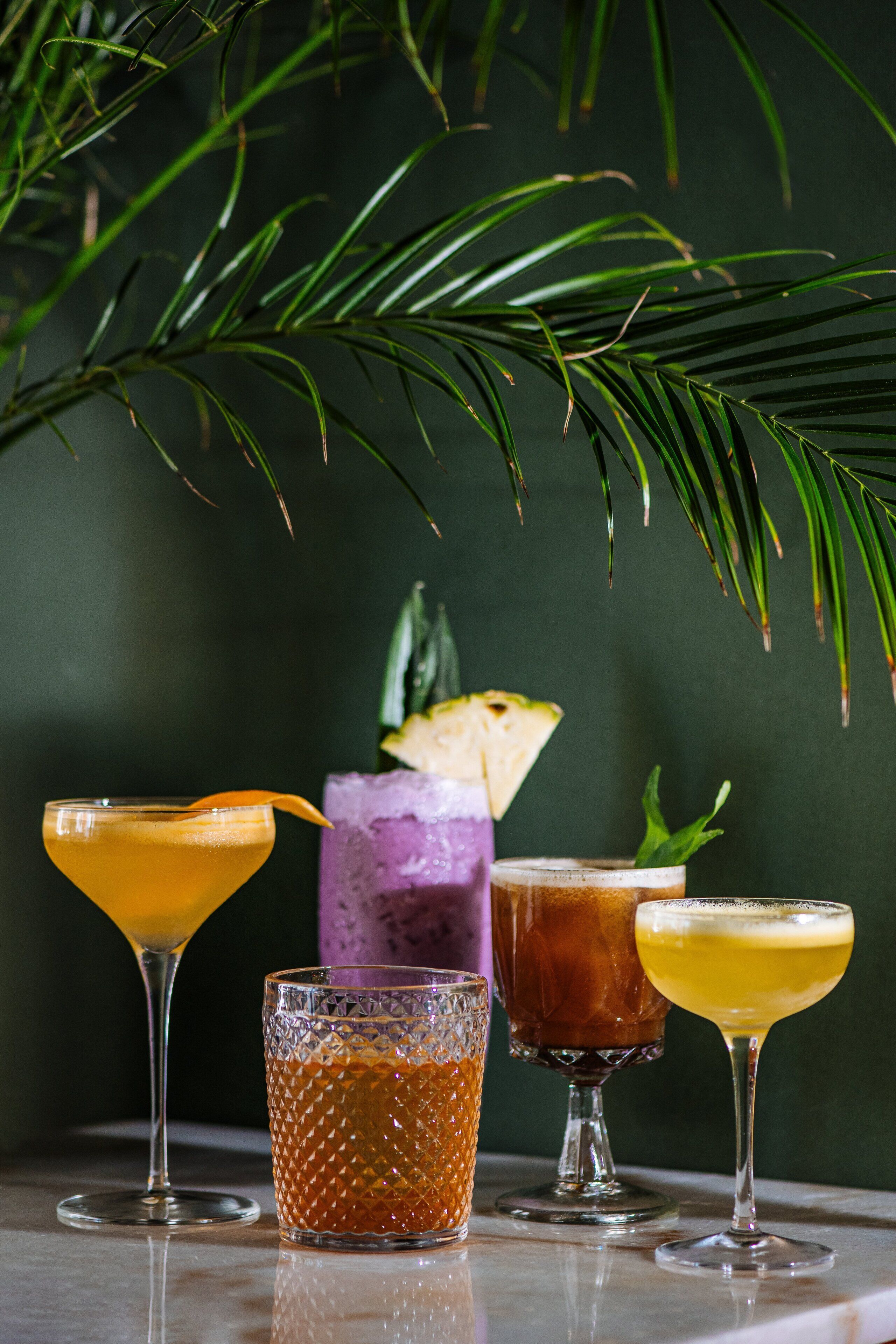 Neljä cocktail-drinkkiä heijastavalla pinnalla, taustalla tumma väri ja palmunlehti.