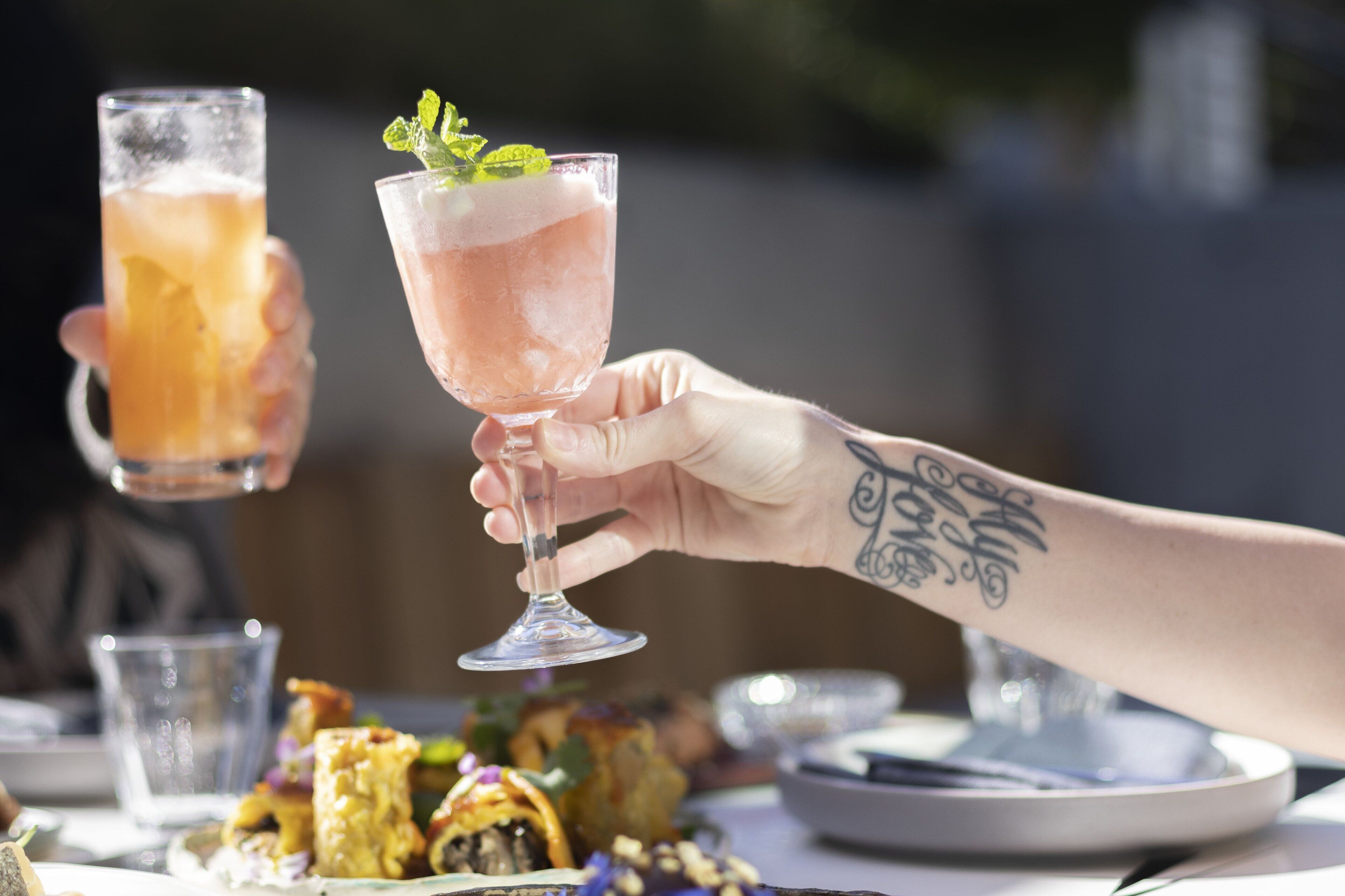 A person holding a glass with a pink drink, another glass with a yellow drink, and a plate with food in the background.