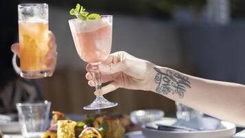 Una persona sosteniendo un vaso con una bebida rosa, otro vaso con una bebida amarilla, y un plato con comida al fondo.