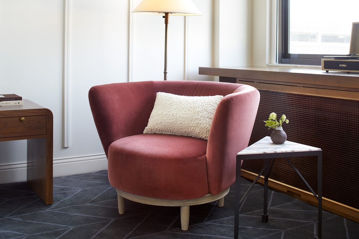 A red armchair with a white cushion, a small table with a vase, and a floor lamp in a room with a radiator and a window.