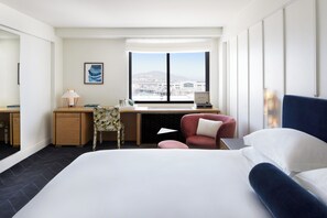 Une chambre d’hôtel avec un grand lit, un bureau avec un ordinateur, une chaise et une vue sur le paysage urbain par une grande fenêtre.
