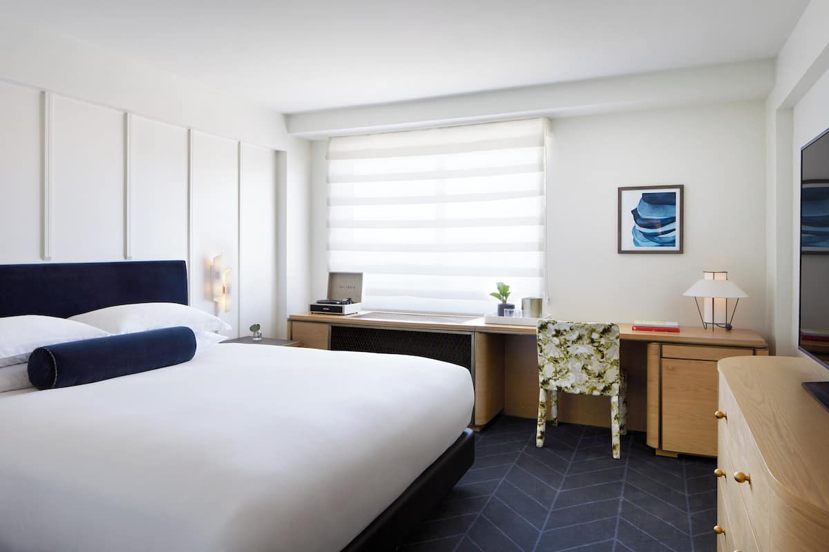 A hotel room with a large bed, a desk, a chair, a small table with a plant, and a window with blinds.