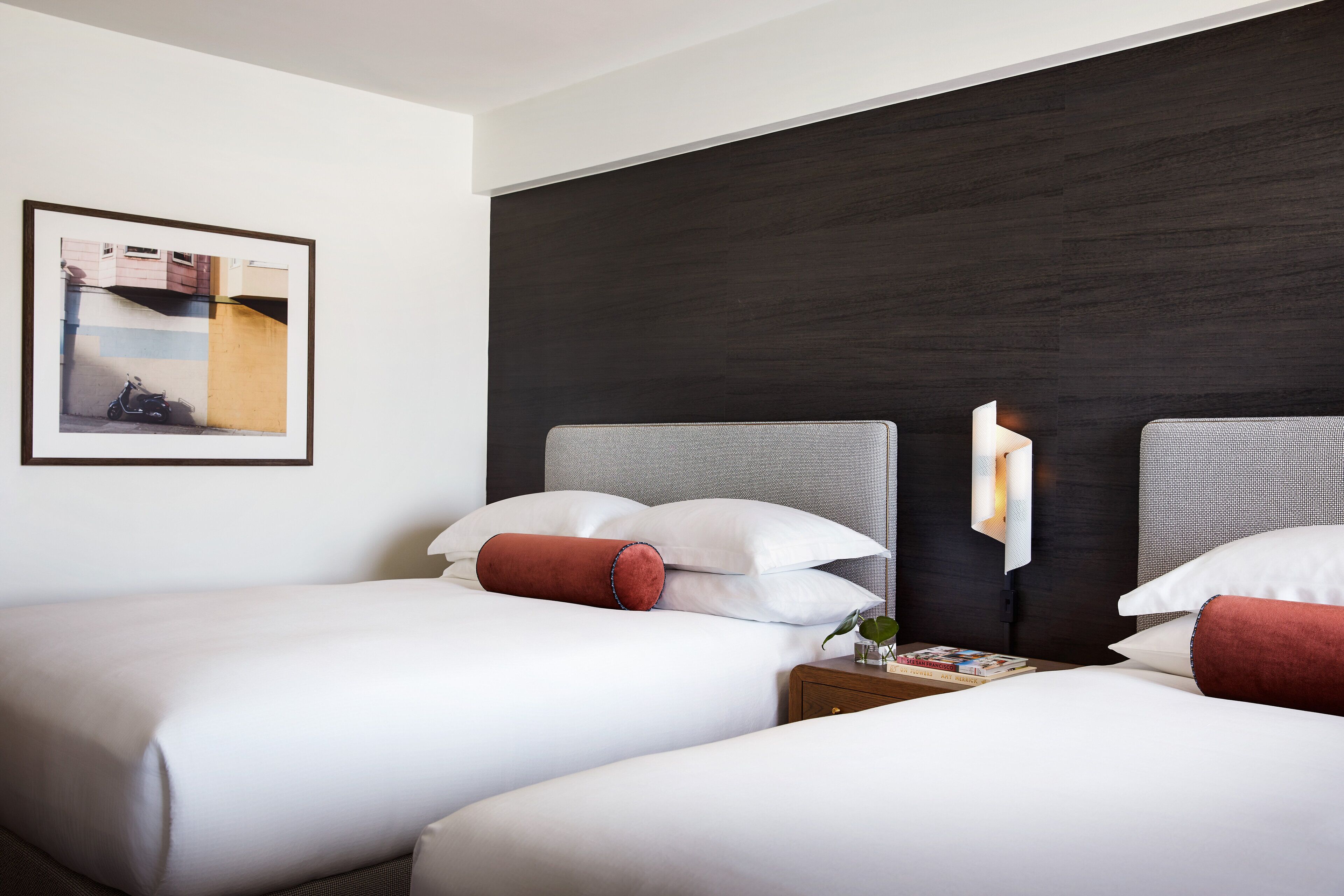 A hotel room with two beds, a dark wood headboard, a framed picture on the wall, and a bedside table with a lamp and books.