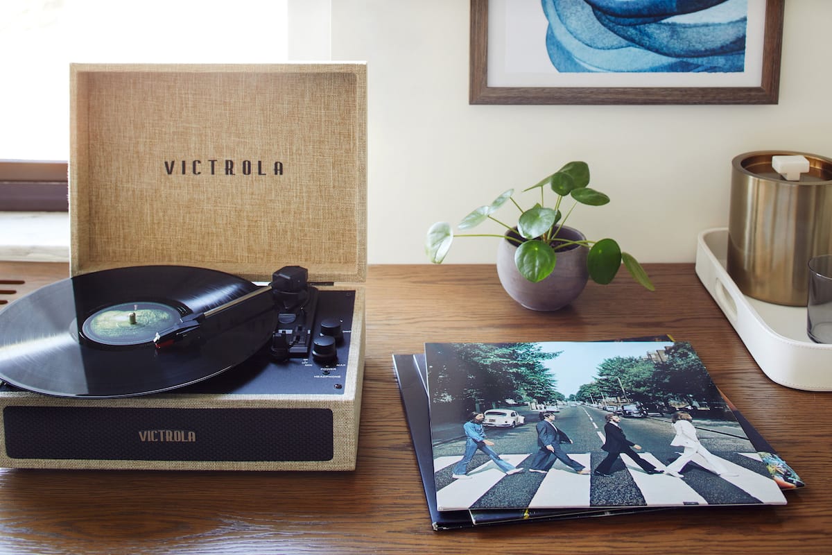 A turntable with a vinyl record, a vinyl record box, a framed picture, a potted plant, and a candle holder on a wooden table.