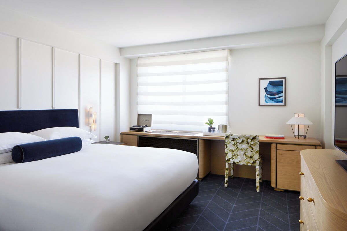 A hotel room with a large bed, a desk, a chair, a small table with a plant, and a window with blinds.