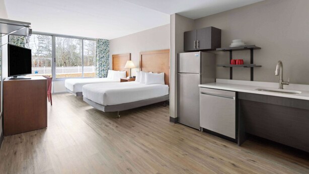 A hotel room with two beds, a TV, a desk, a refrigerator, and a kitchen area with a sink and shelves.