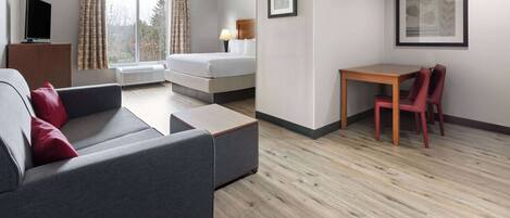 Ein modernes Hotelzimmer mit grauem Sofa, einem Holztisch mit zwei Stühlen, einem Bett mit weißer Bettwäsche, einem Flachbildfernseher auf einer Holzkommode und einem großen Fenster mit Vorhängen.