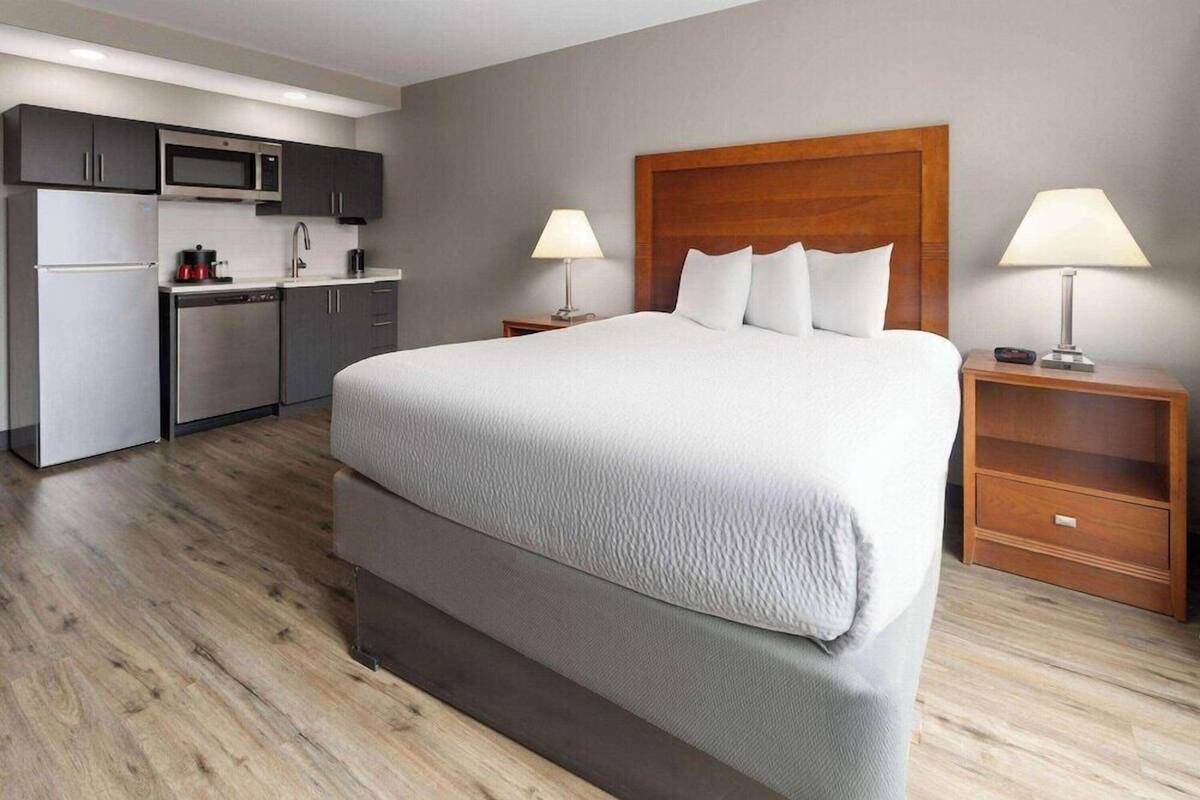 A hotel room with a large bed, a wooden headboard, a kitchen area, and two bedside lamps.