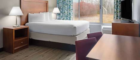 A hotel room with a bed, two bedside lamps, a TV on a wooden stand, a wooden table, and a window with curtains.