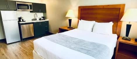 A hotel room with a bed, two bedside tables with lamps, a wooden headboard, a kitchenette with a microwave, and a refrigerator.