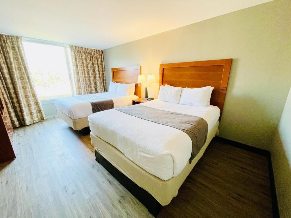 A hotel room with two beds, a wooden headboard, a nightstand with a lamp, and a window with curtains.