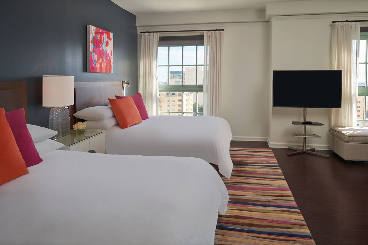 A hotel room with two beds, a TV, a window with curtains, and a view of buildings.