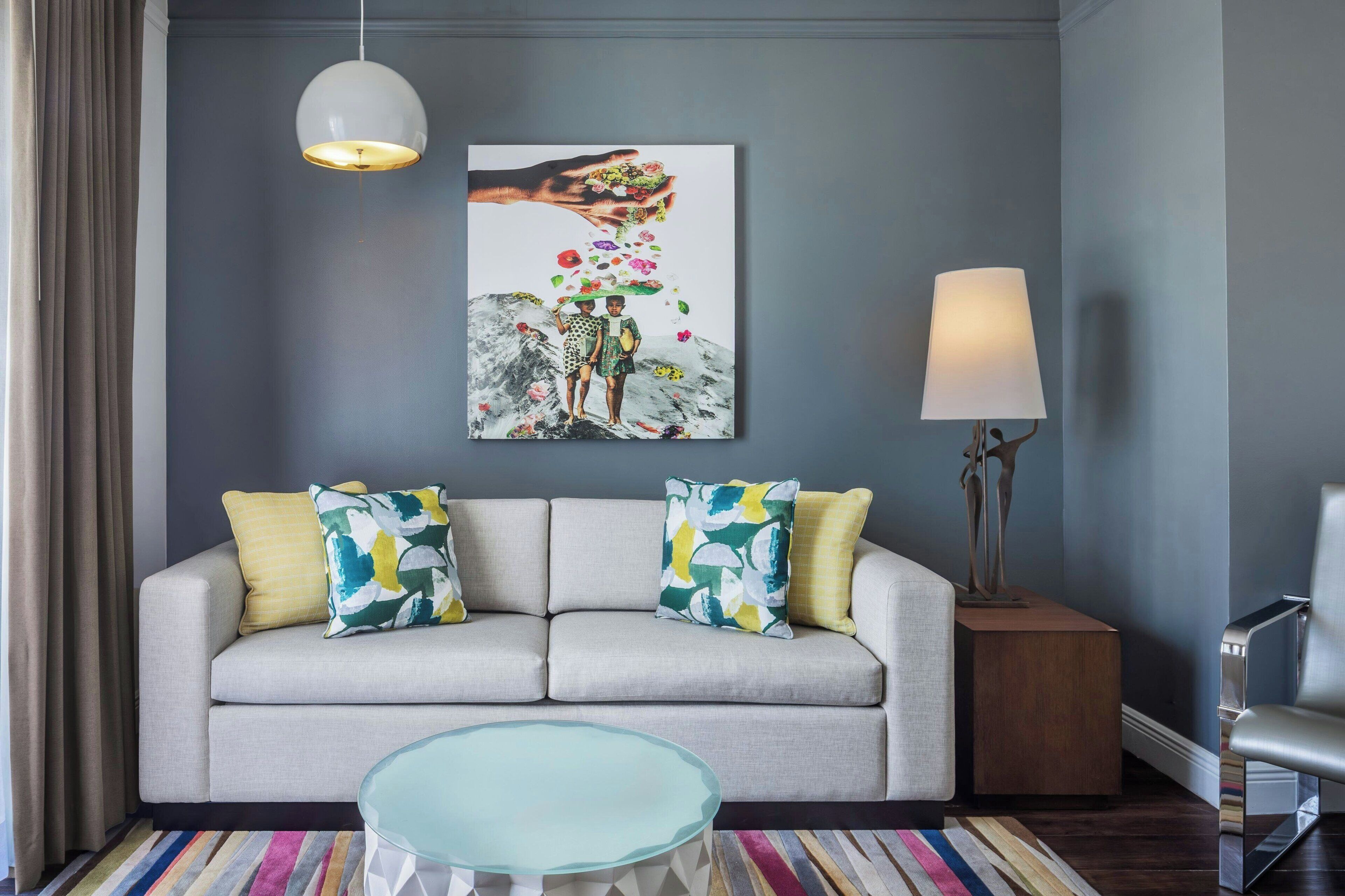 Een moderne woonkamer met een grijze bank, kleurrijke kussens, een glazen salontafel, een schilderij aan de muur en een vloerlamp.