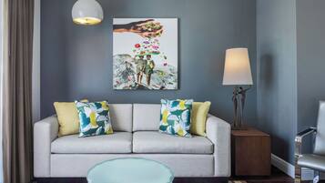 Een moderne woonkamer met een grijze bank, kleurrijke kussens, een glazen salontafel, een schilderij aan de muur en een vloerlamp.