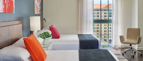 Habitación de hotel con dos camas, un escritorio, una silla, un ventanal con vistas a edificios y obras de arte en las paredes.