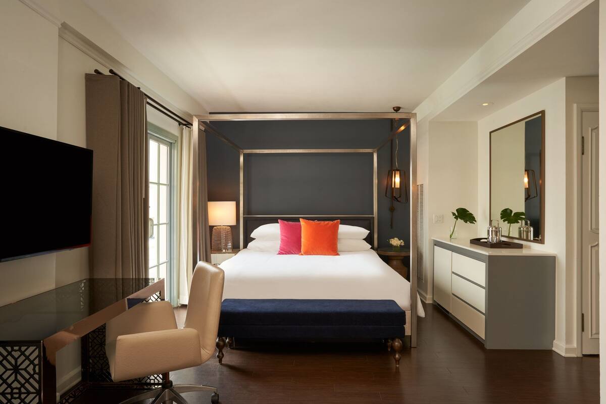 A hotel room with a large bed, a desk with a chair, a TV, a bedside table with a lamp, and a mirror.