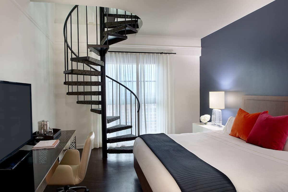 A modern hotel room with a curved black spiral staircase, a large bed with a black headboard, and a desk with a chair.