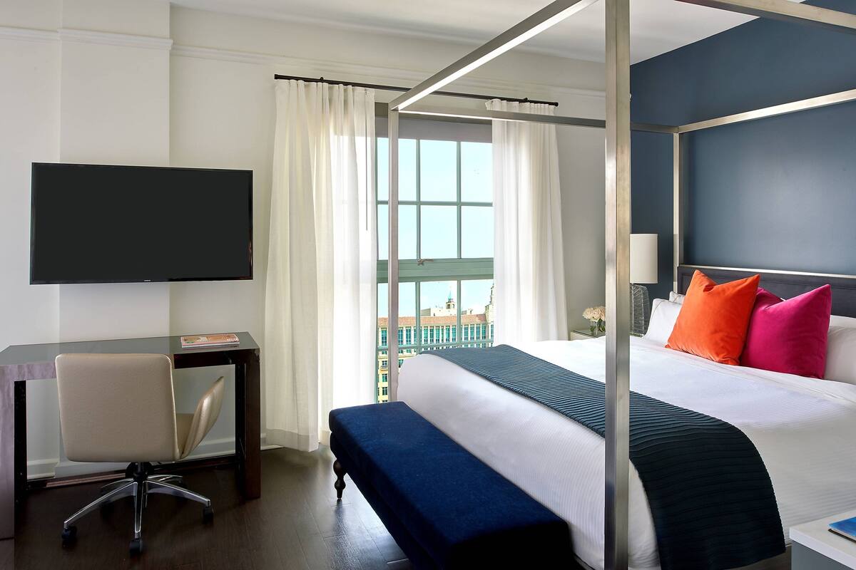 A hotel room with a four-poster bed, a desk with a chair, a television, and a view of the city through large windows.