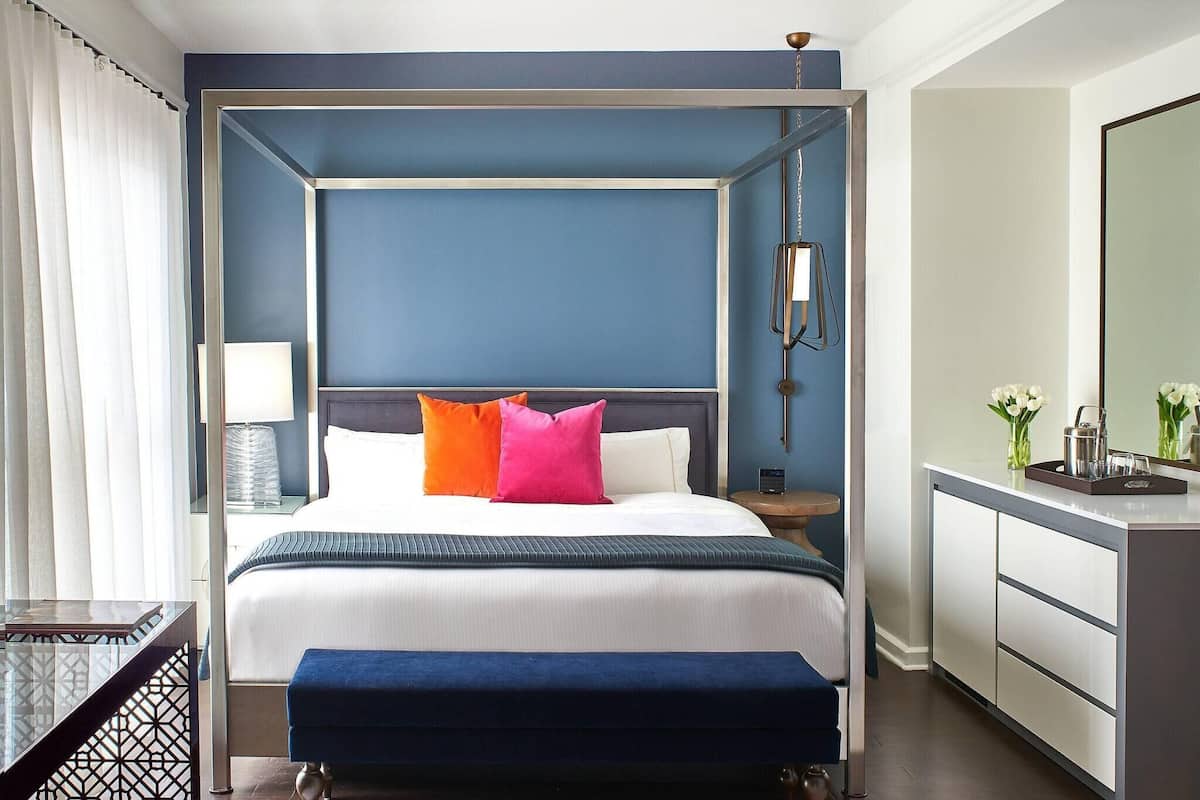 A hotel room with a four-poster bed, bedside tables, a bench, a nightstand, a mirror, and a dresser.