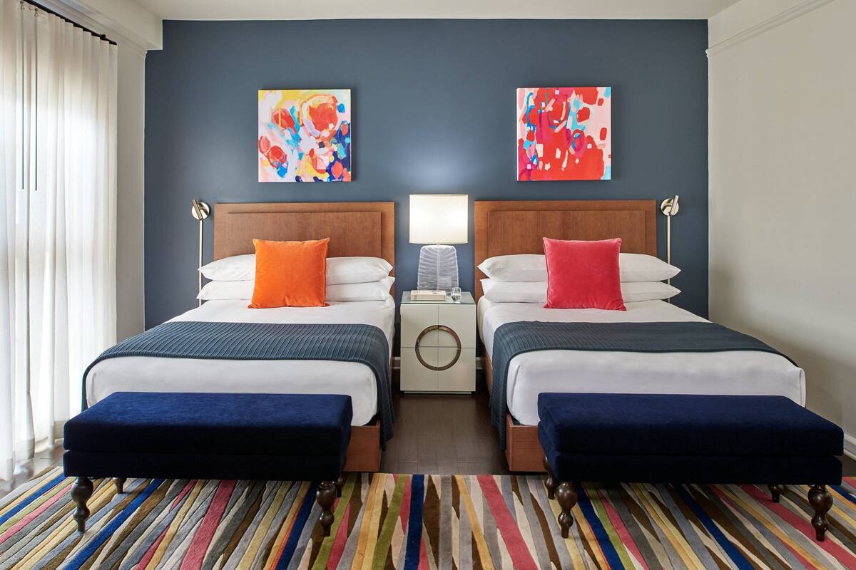 A hotel room with two beds, a nightstand, a lamp, and colorful artwork on the walls.