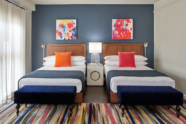 A hotel room with two beds, a nightstand, a lamp, and colorful artwork on the walls.