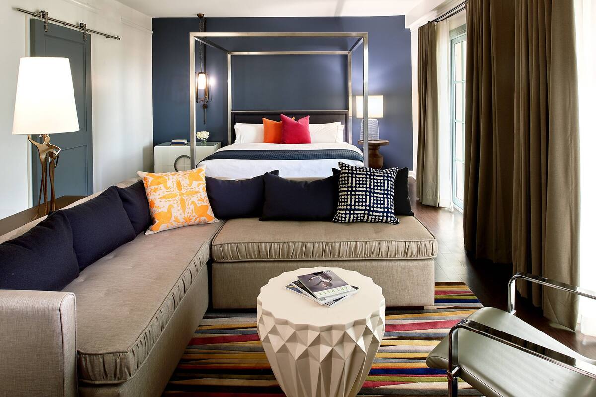 A modern hotel room with a four-poster bed, a sofa, a small round table, and a bench.