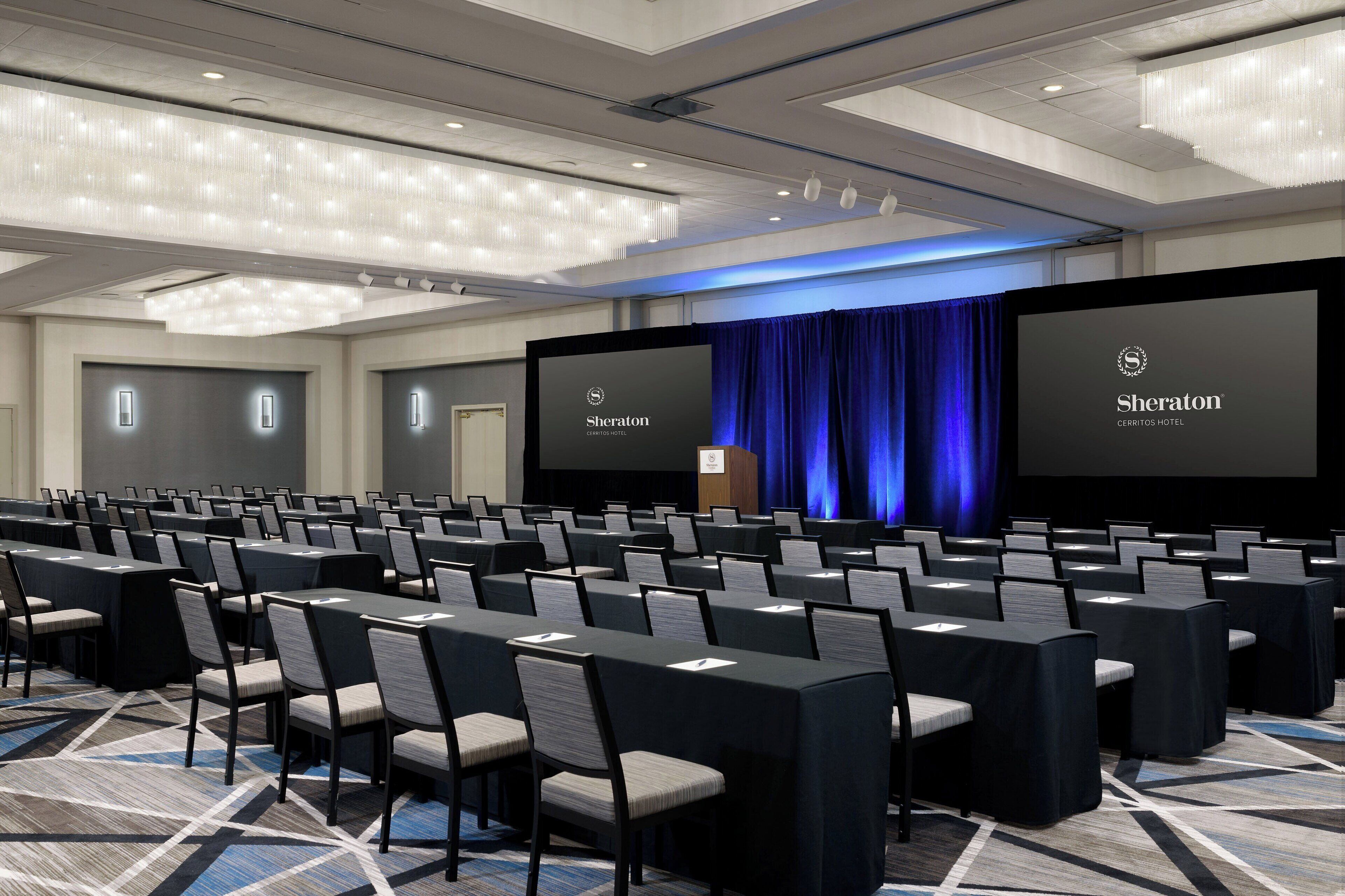 Un salón de conferencias con filas de mesas y sillas, un escenario con un telón azul y dos pantallas grandes mostrando el logotipo de Sheraton.