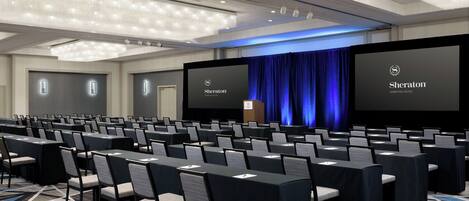 Un salón de conferencias con filas de mesas y sillas, un escenario con un telón azul y dos pantallas grandes mostrando el logotipo de Sheraton.