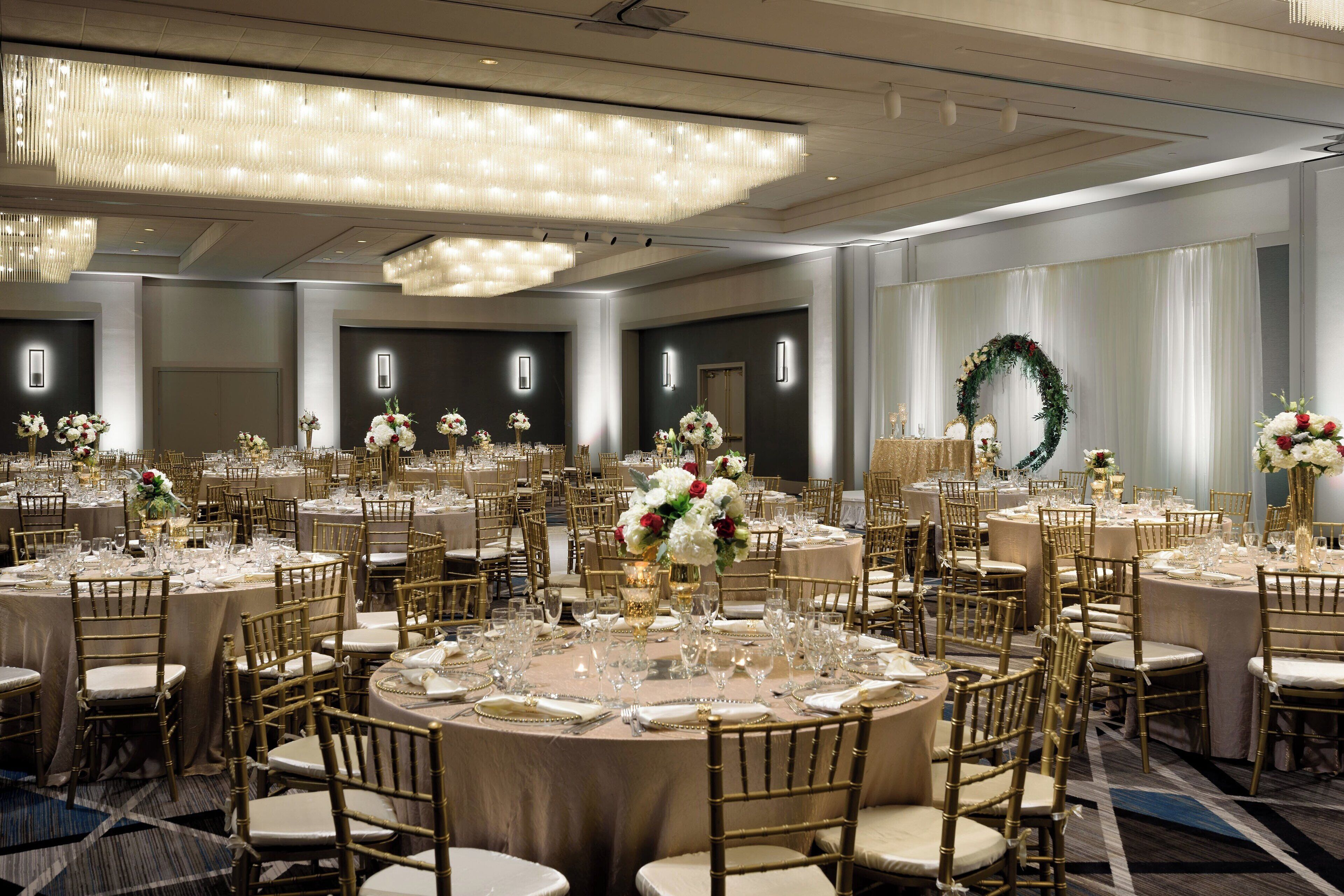 Ein Bankettsaal mit runden Tischen, goldenen Stühlen und floralen Tischdekorationen.
