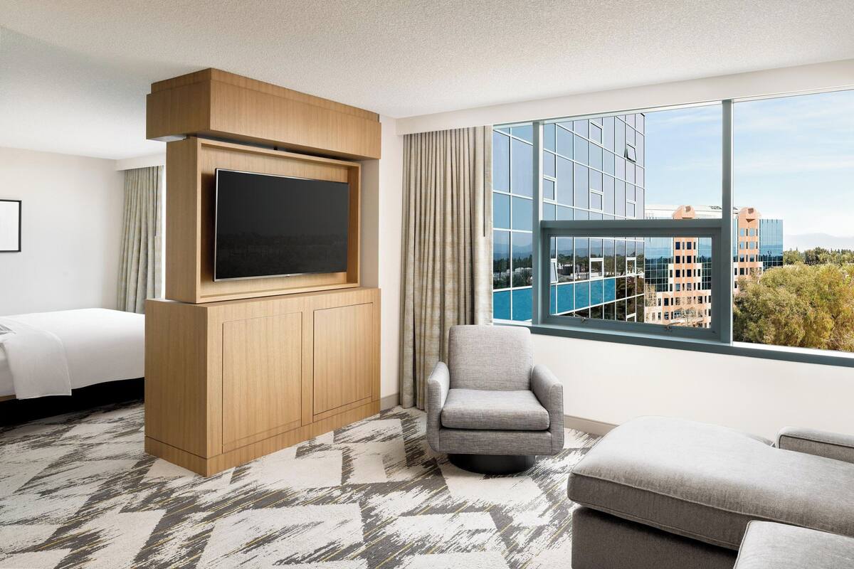 Habitación de hotel con televisor grande, sillón cómodo, sofá y vista de la ciudad a través de amplias ventanas.