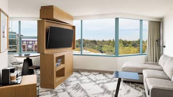 A modern hotel room with a large TV, a sofa, a coffee table, and a dining area with chairs and a table.