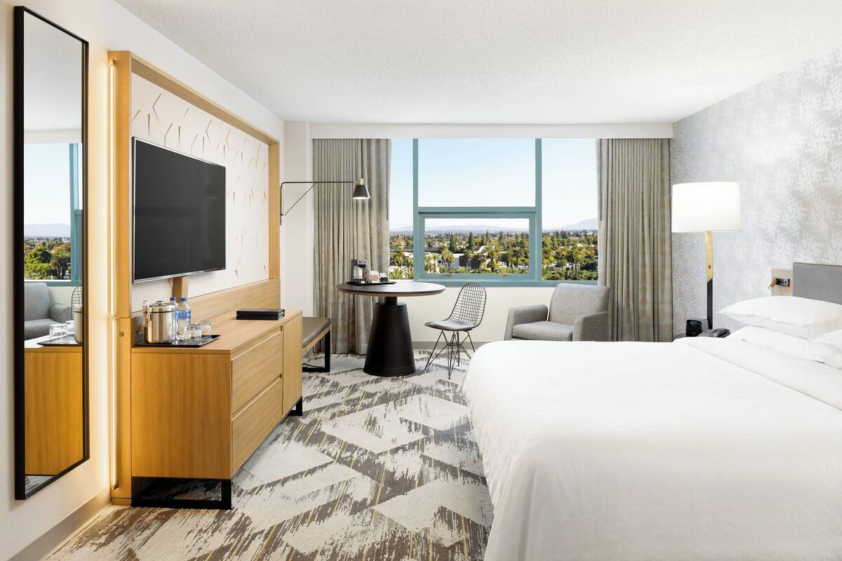Habitación de hotel con una cama grande, televisión, escritorio con silla, una mesita y vista a la ciudad.